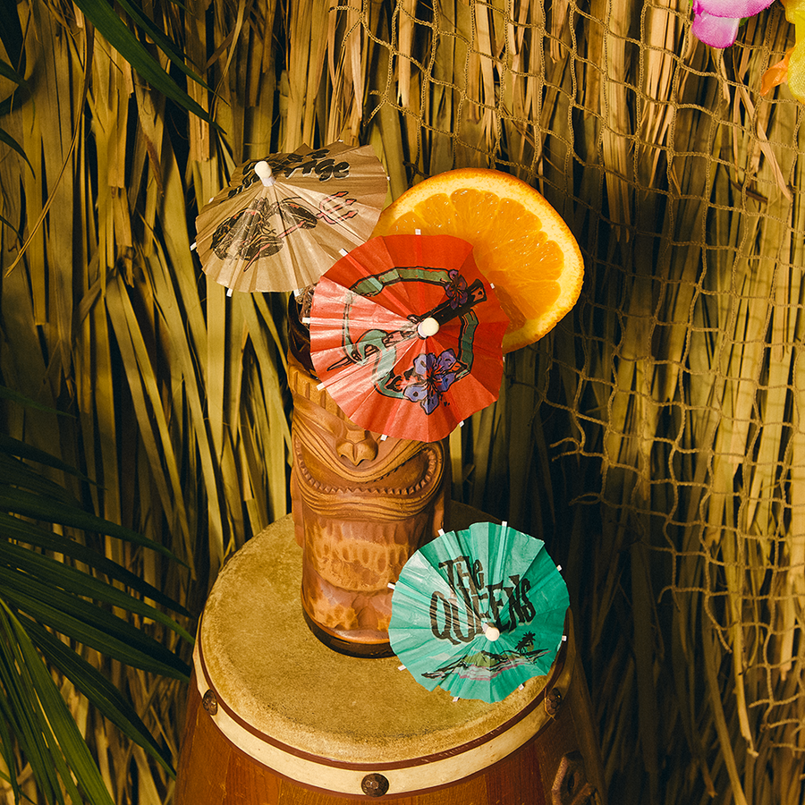 Tiki mug with cocktail umbrellas and an orange slice on a barrel against a thatched wall.
