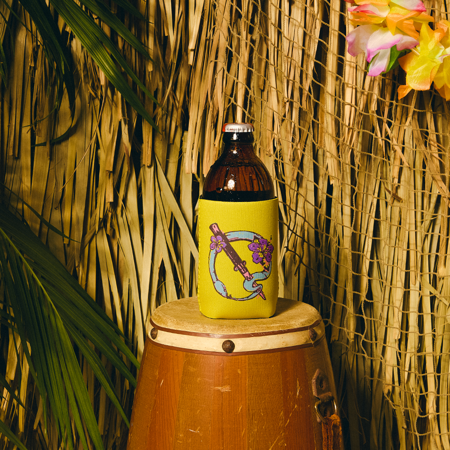 Bottle with a colorful label on a yellow koozie, placed on a wooden barrel against a thatched wall.